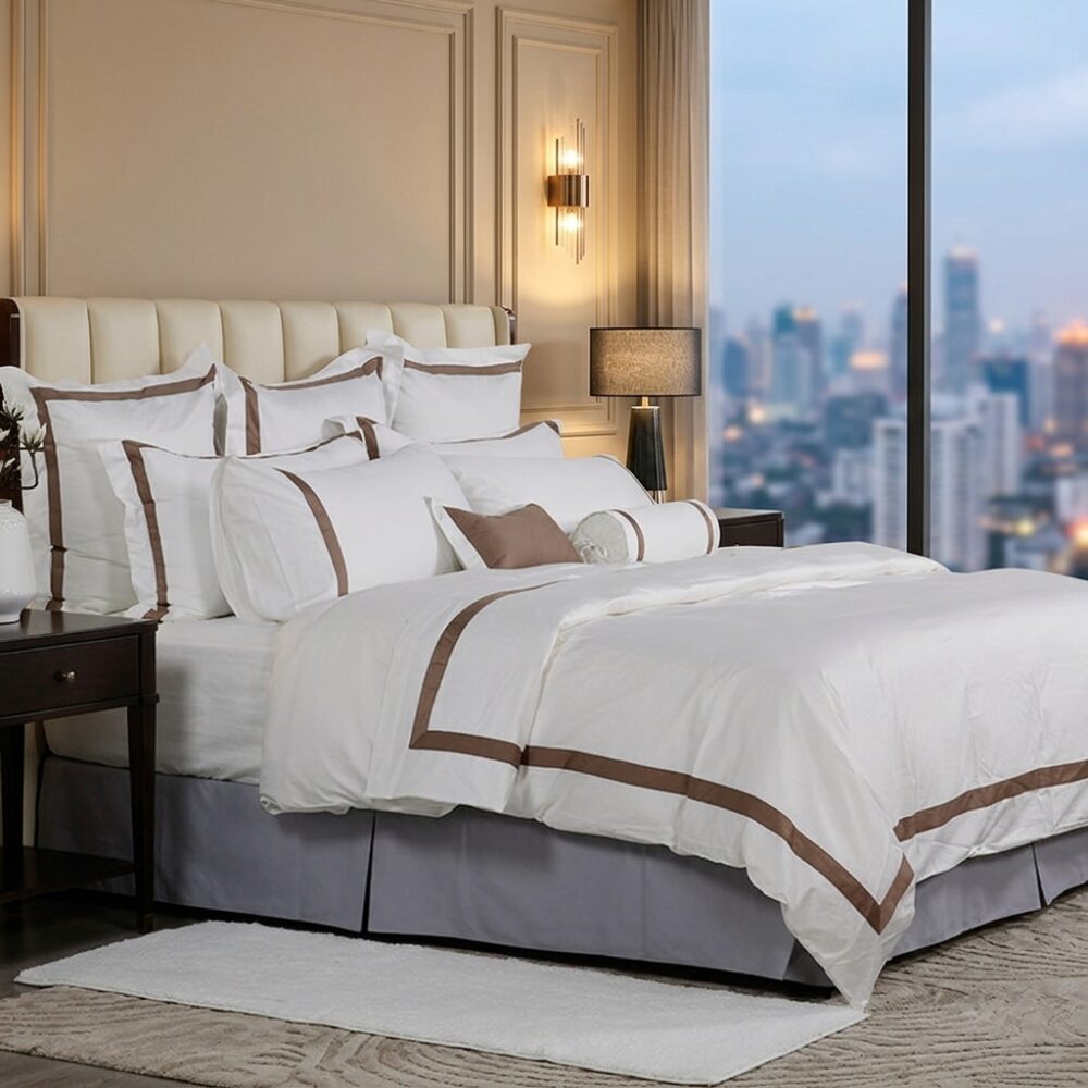 A luxury hotel bed with white linens featuring bronze-brown border embroidery and a view of a city skyline at dusk.
