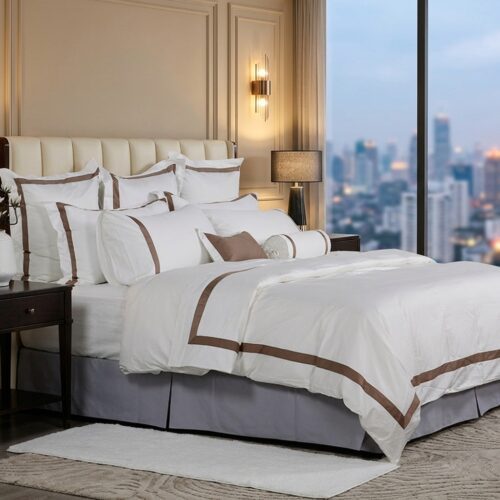 A luxury hotel bed with white linens featuring bronze-brown border embroidery and a view of a city skyline at dusk.