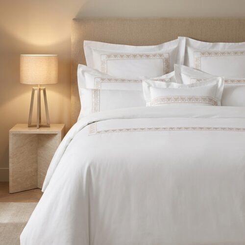 Luxury hotel bed featuring white cotton linens with champagne gold Greek key embroidery against a beige tufted headboard.