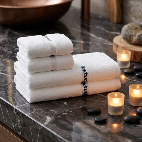 Set of four folded luxury white cotton hotel towels with navy blue script embroidery displayed on a dark marble surface surrounded by glowing candles and spa stones
