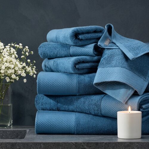 Stack of luxury steel blue cotton towels in various sizes folded and styled with white flowers and a lit candle on a dark stone surface