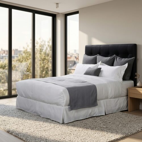 A modern hotel bed featuring crisp white linens, a slate gray bed runner, and charcoal gray pillows in a bright room with large windows.