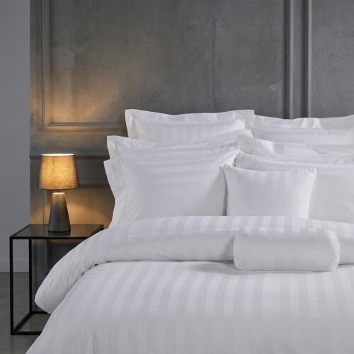 A luxury hotel bed featuring white satin-striped linens and a matching bolster pillow against a grey concrete-textured wall.