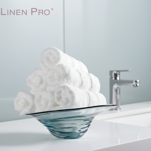 A neat pyramid of six rolled white luxury hotel washcloths displayed in a modern translucent blue glass bowl on a white bathroom vanity.