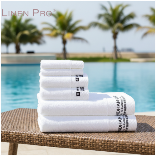 A stack of plush white hotel towels featuring black custom embroidered logos and textured borders on a wicker lounge chair by a swimming pool.
