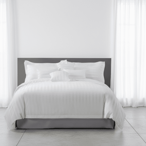 A luxury hotel bed featuring white satin-striped linens, multiple layered pillows, and a tailored grey bed skirt against a grey headboard.
