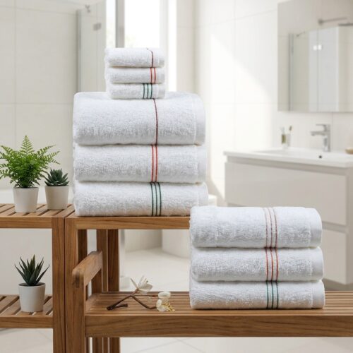A stacked collection of luxury plush white hotel bath towels and washcloths with thin red, orange, and green embroidered accent stripes on a wooden spa shelf.