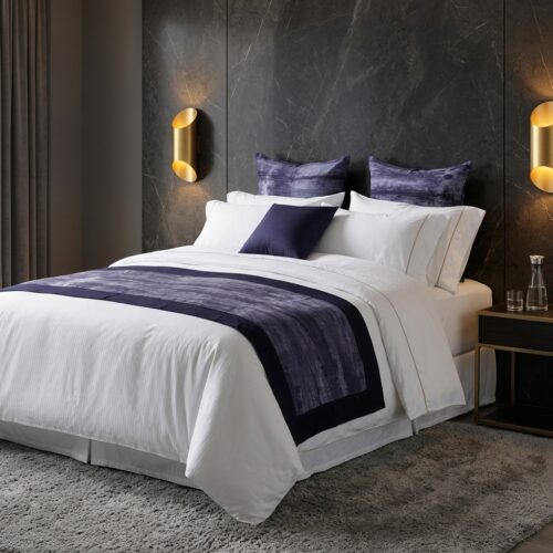 A luxury boutique hotel king bed dressed in white sateen stripe sheets with an abstract dark violet bed runner and decorative throw pillows against a black marble wall.