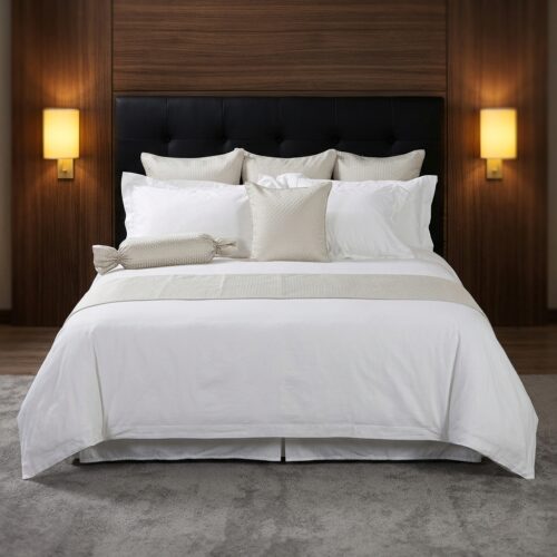A luxury hotel king bed dressed in crisp white bedding with a textured champagne gold bed runner, a matching accent cushion, and a candy-wrap bolster pillow against a wood panel headboard.