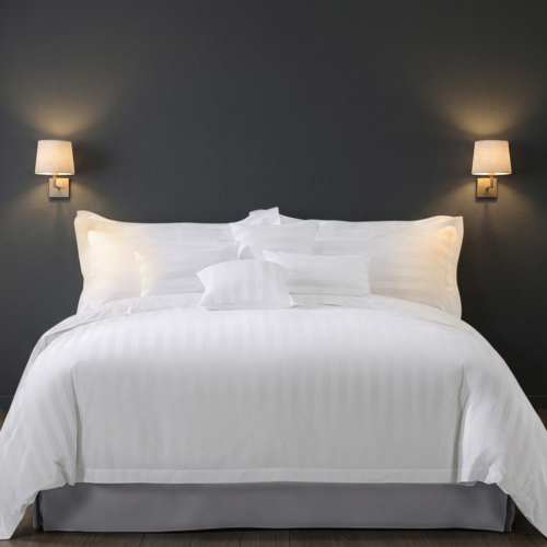 Luxury white satin stripe hotel bedding set with layered pillow arrangement on a king bed flanked by warm wall sconces against a charcoal gray feature wall in a modern hotel room