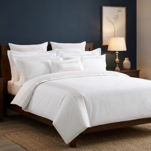 A luxury boutique hotel king bed dressed in a white bedding set with champagne honeycomb geometric embroidery detail against a deep navy blue accent wall.