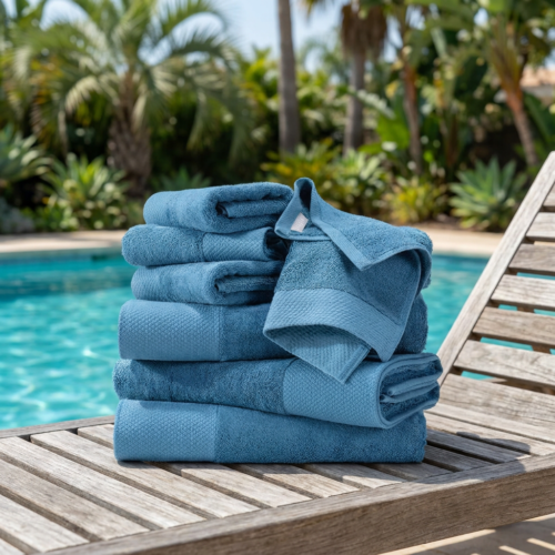 Stack of luxury teal blue cotton resort pool towels in multiple sizes folded and displayed on a wooden sun lounger beside a blue swimming pool with tropical palms in the background