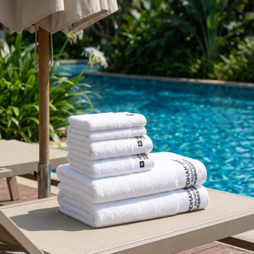 Set of six luxury white hotel pool towels with black logo embroidery neatly stacked on a beige sun lounger beside a tropical resort swimming pool under a poolside umbrella