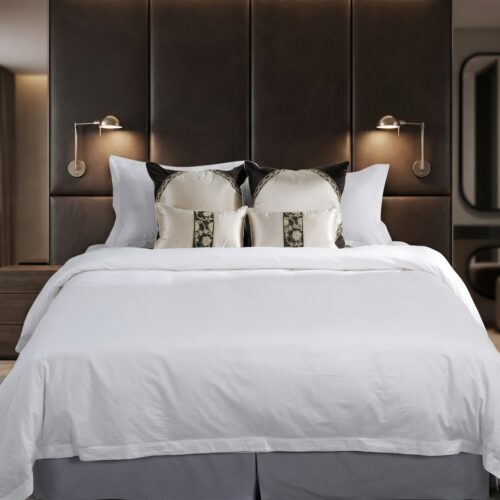 A luxury hotel bed dressed in crisp white linens with decorative embroidered jacquard accent pillows against a tall dark leather headboard panel.