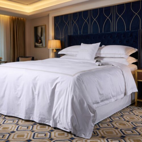 A luxury hotel king bed dressed in crisp white linens with a tan embroidered border duvet cover and a dark blue tufted headboard.