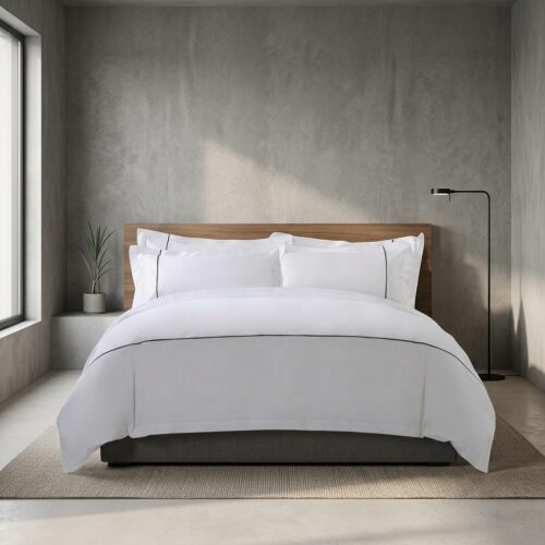 A minimalist hotel king bed dressed in crisp white sateen linens with a black embroidered line duvet cover against a raw concrete wall.
