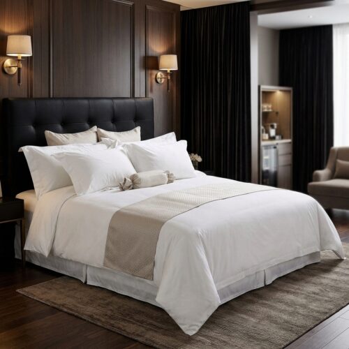 A luxury hotel king bed dressed in crisp white linens, featuring a champagne gold bed runner and a matching candy-wrapper style bolster pillow against a tufted black headboard.