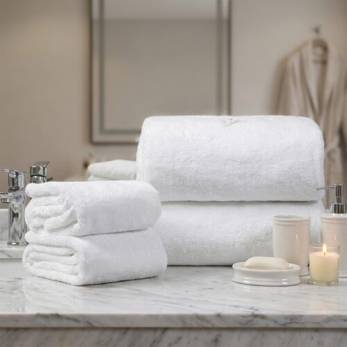 A stacked set of fluffy white hotel bath towels and folded hand towels arranged on a marble bathroom counter next to a soap dish and lit candle.