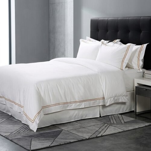 A luxury hotel king bed dressed in crisp white satin linens with a gold double-line embroidered duvet cover and a black tufted headboard.
