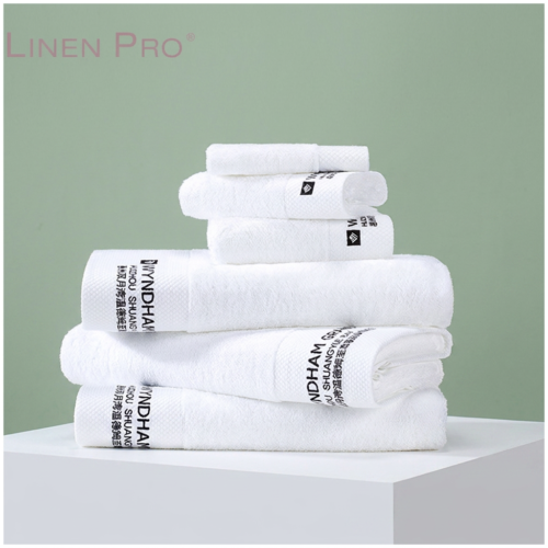 A neat stack of luxury white hotel bath towels, hand towels, and washcloths featuring custom black brand embroidery on a white block against a sage green background.