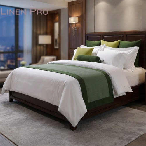 A luxury hotel king bed dressed in crisp white bedding with a two-tone moss green bed runner, matching green pillows, and a cylindrical bolster cushion against a dark wood headboard.