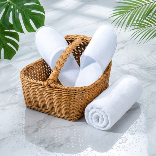 A pair of luxury white closed-toe velvet slippers next to a neatly rolled white cotton terry towel on a wet marble surface with tropical green leaves.