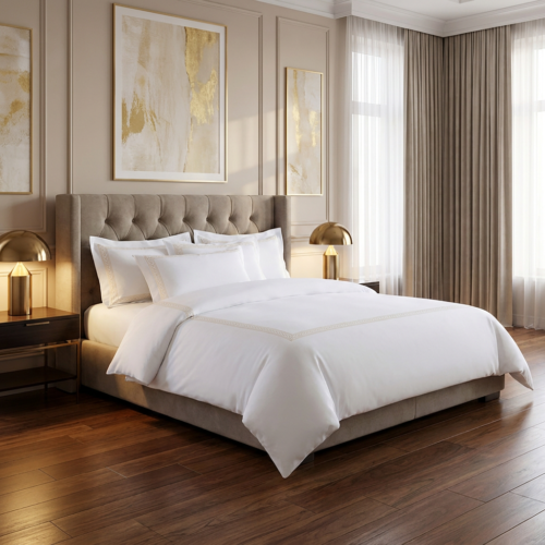 Luxury white hotel bedding set with gold trim border detail on a king bed with a grey tufted velvet headboard flanked by brass dome table lamps in an elegant beige and gold hotel suite with abstract gold artwork above