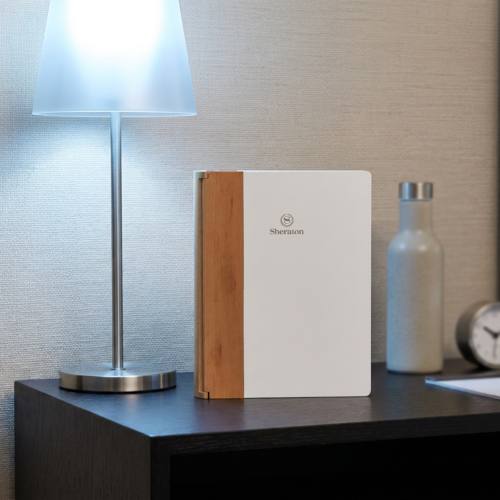 A modern rectangular matte white hotel guest directory binder with a natural wood spine standing on a dark wooden desk under a glowing lamp.