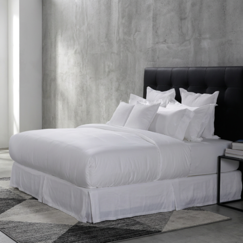 Premium white hotel duvet cover, stacked Oxford pillows, and matching bed skirt styled against a black tufted leather headboard and concrete wall.
