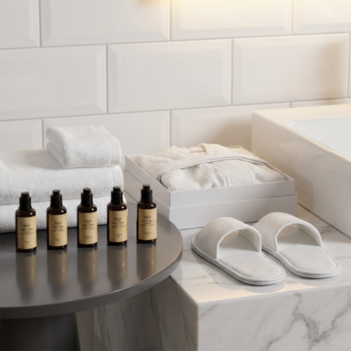 A comprehensive display of premium white hotel towels, a boxed white bathrobe, plush slippers, and five amber cosmetic pump bottles on a bathroom vanity.