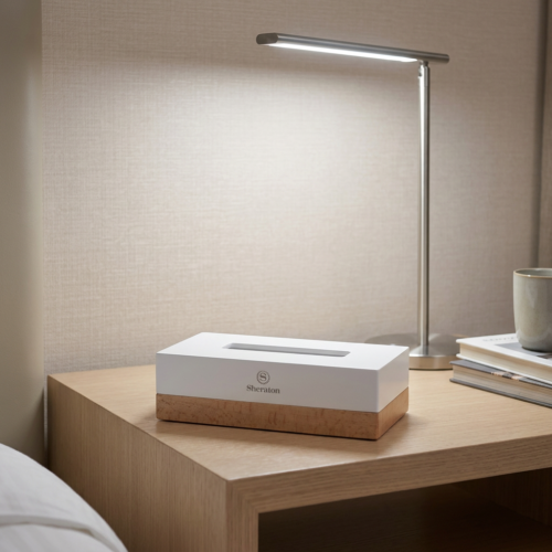 A modern rectangular matte white tissue box cover with a natural wood base sitting on a hotel room wooden nightstand beneath a glowing metal desk lamp.
