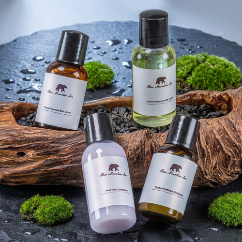 A collection of four mini hotel toiletry bottles including shampoo and conditioner arranged over a rustic wooden log and black stones.