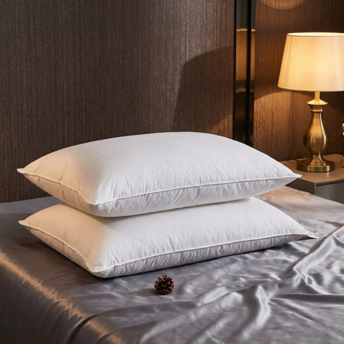 Two stacked plush white hotel sleeping pillows with piped edges resting on a silky grey sheet beside a glowing warm table lamp.