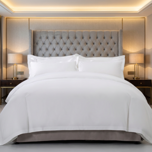 Premium pure white hotel duvet cover set with layered Oxford pillowcases on a tufted grey headboard bed flanked by warm glowing nightstand lamps.