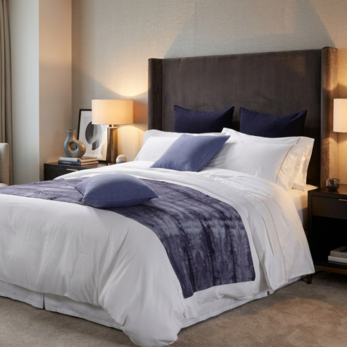 Galaxy premium white hotel bedding layered with a textured indigo blue runner and accent pillows against a tall dark upholstered headboard.