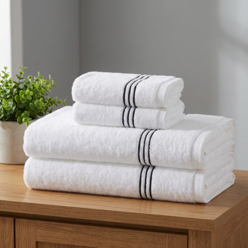 A neatly stacked set of white hotel towels featuring three woven black accent stripes, sitting on a wooden vanity counter next to a potted green plant.