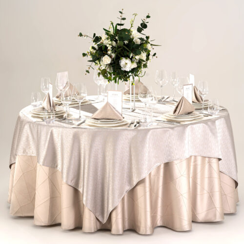 Hotel round banquet table styled with a champagne geometric skirt, silver overlay, stitched placemats, folded napkins, and green floral centerpiece.