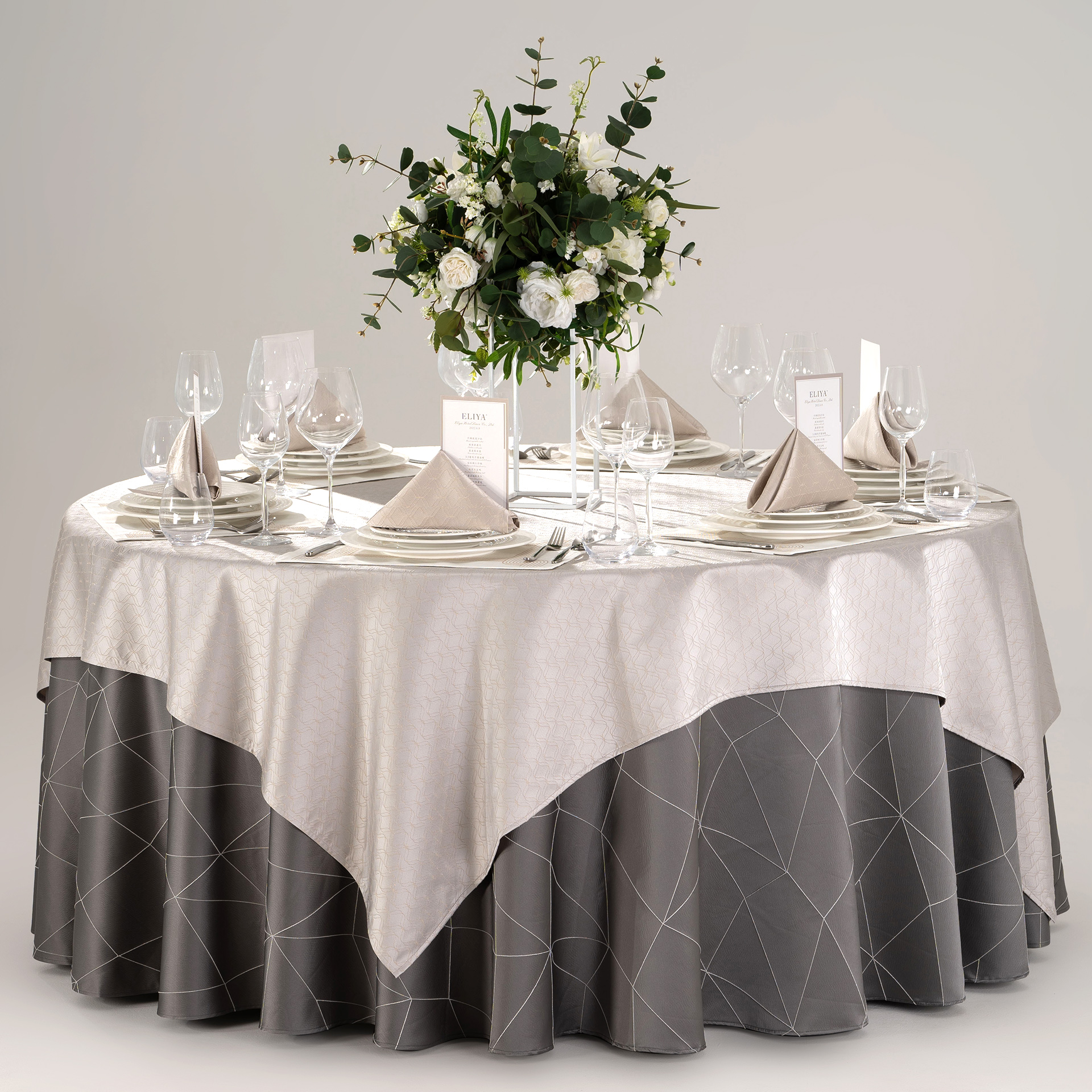 Hotel round banquet table styled with a dark grey geometric skirt, silver overlay, stitched placemats, folded napkins, and green floral centerpiece.