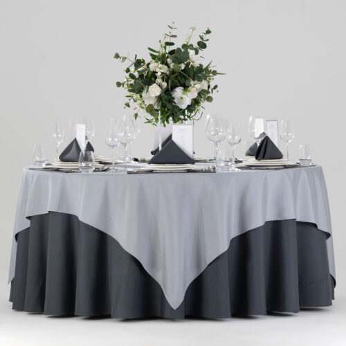 Hotel round banquet table styled with a charcoal base skirt, silver gray square overlay, dark placemats, folded napkins, and white floral centerpiece.