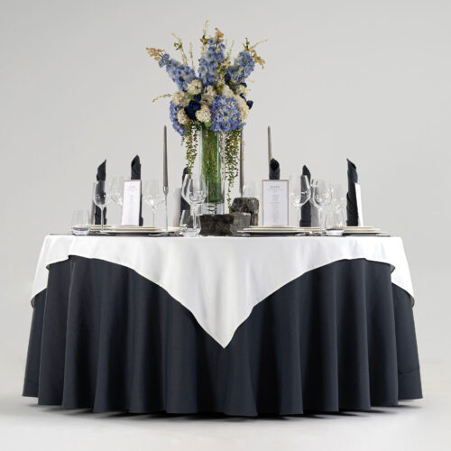 Hotel round banquet table styled with a black base skirt, white square overlay, gold embroidered placemats, folded napkins, and blue floral centerpiece.