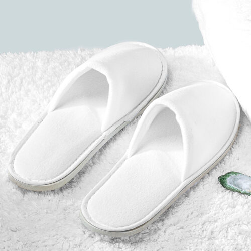 Pair of white open-toe fleece hotel slippers with cushioned insoles resting on a plush rug.