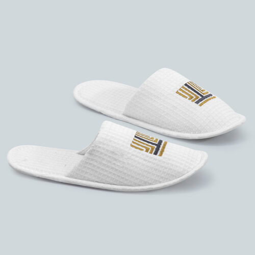 Pair of white waffle weave closed-toe hotel slippers with custom gold and navy embroidered logo.